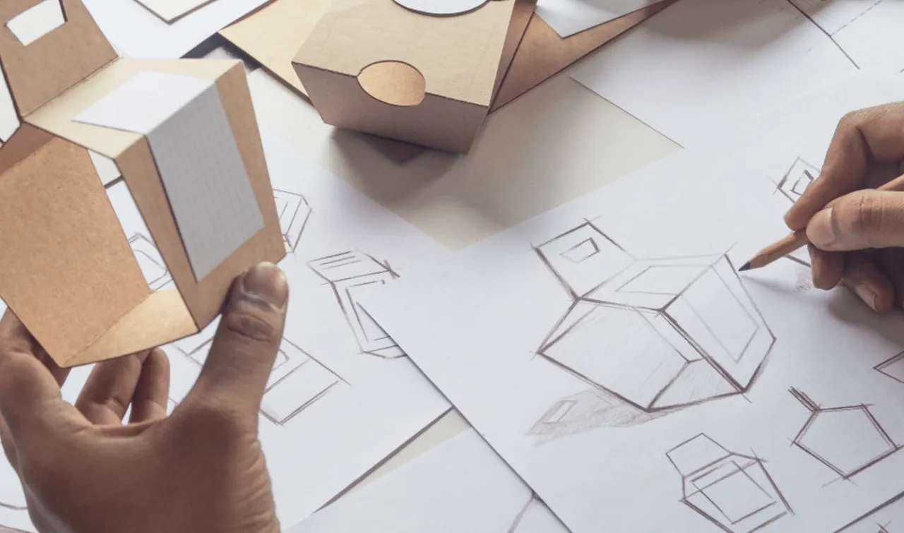 Mão desenhando a planificação de uma caixa ao lado de protótipos de papelão